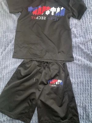 Svart set från Trapstar med tryck - Säljer ett svart set från Trapstar med färgglatt tryck på både t-shirt och shorts. Trycket har röd, vit och blå text med 'Trapstar It's a Secret'. Setet är i mjukt material och har avslappnad passform. Perfekt för en chill och trendig look.
