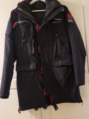 Svart parkas med röda detaljer MA.59 - Snygg svart parkas från MA.59 med röda detaljer och flera praktiska fickor. Jackan har quiltat innerfoder, hög krage och dragkedja med tryckknappar. Perfekt för kalla dagar och har en modern, sportig look.