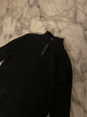 Svart stickad tröja med halv dragkedja - Mysig svart stickad tröja med hög krage och halv dragkedja framtill. Tröjan har lång ärm och ribbstickad struktur som ger en snygg och enkel look. Perfekt för lager på lager och passar till det mesta i garderoben.