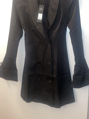 Svart kavajklänning med vida ärmar - Stilren svart kavajklänning med dubbelknäppning och breda slag. Klänningen har långa, vida ärmar och en figurnära passform. Perfekt för dig som vill ha en elegant och trendig look.