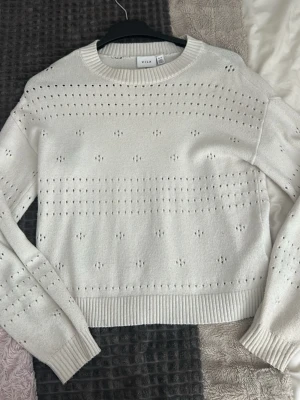 Vit stickad tröja från VILA - Snygg vit stickad tröja från VILA med hålmönster över hela tröjan. Tröjan har rund halsringning, långa ärmar och ribbade muddar vid ärmslut och nederkant. Perfekt för lager på lager och enkel att matcha med jeans eller kjol. Det har blivit ett hål på baksidan vid halsringningen, inget man direkt tänker på men går säkert att sy upp