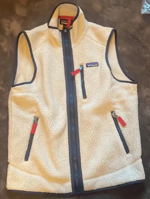 fleeceväst från Patagonia - Mysig beige fleeceväst från Patagonia med marinblå detaljer och dragkedjor med röda dragare. Västen har hög krage, tre fickor med dragkedja och är tillverkad i mjukt teddyfleece-material. Perfekt för lager-på-lager och outdoor vibes.