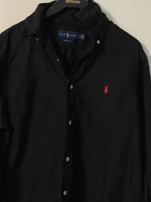 Svart skjorta från Ralph Lauren - Klassisk svart skjorta från Ralph Lauren med röd broderad logga på bröstet. Skjortan har knappar framtill och en tidlös design som passar till mycket. Tillverkad i mjuk bomull och har lång ärm. Knappt använd och nypris 1800. 