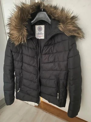 Svart pufferjacka från Saint Vincent - Snygg svart pufferjacka från Saint Vincent med huva och dragkedja framtill. Jackan har två praktiska fickor med dragkedja och quiltad design som håller värmen. Perfekt för kalla dagar och enkel att matcha med olika outfits.