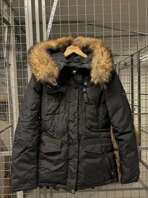 Jacka - Svart vinter jacka från Hollies med stor huva där pälsen går att ta bort. Jackan har flera praktiska fickor, dragkedja och knappar framtill samt. Fickorna vid sidan med dragkedja är även väldigt sköna invändigt. Perfekt för kalla dagar och riktigt snygg till vintern och hösten. Jackan är i mycket fint skick!🤍🤍🤍 nypris: 4500kr