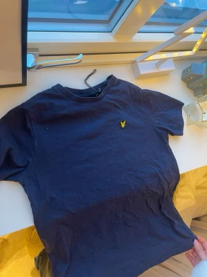 Marinblå t-shirt från Lyle & Scott - Mörkblå t-shirt från Lyle & Scott med klassisk rund hals och korta ärmar. På bröstet sitter den ikoniska gula örnloggan broderad. Perfekt basic-plagg att ha i garderoben. Den är i storlek 12-13 år. Aldrig använd 