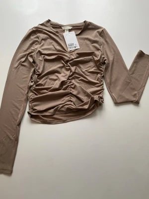 Beige långärmad topp med rynk - Trendig beige långärmad topp från Bik Bok med snygg rynkdetalj framtill och croppad passform. Tillverkad i mjukt och stretchigt material som sitter skönt på kroppen. Perfekt för dig som gillar stilrena plagg med en twist.