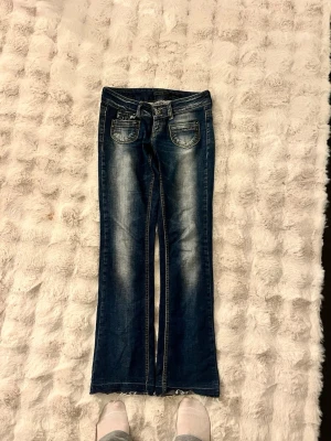 Blå bootcut jeans med unikt märke  - Säljer ett par blå bootcut jeans med snygga slitningar och tvättade detaljer. Jeansen har fem fickor, låg midja och råa kanter vid bensluten. Perfekta för en avslappnad och trendig look.