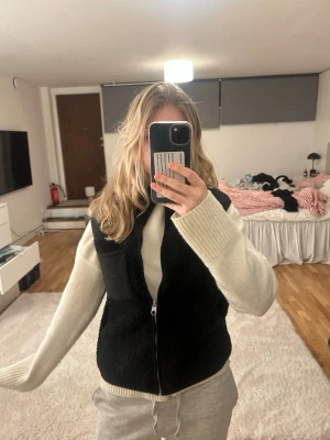 Svart teddyväst från Pieces XS - Svart teddyväst från Pieces i storlek XS. Västen har en hög krage, dragkedja framtill och en liten ficka på bröstet. Materialet är mjukt och fluffigt, perfekt för lager-på-lager. Enkel och stilren design som funkar till många outfits.