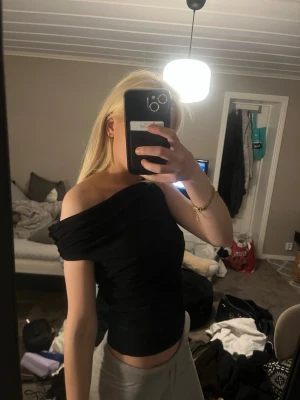 Svart offshoulder topp - Trendig svart offshoulder topp med tight passform och draperad effekt. Toppen är ärmlös och har en asymmetrisk skärning som ger en cool och modern look. Perfekt att styla med både jeans och kjol för en snygg outfit.