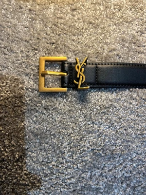 Svart YSL bälte i skinn med gulddetaljer - Stilrent svart bälte från Yves Saint Laurent i skinn med klassiskt YSL-logo i guld och matchande guldfärgat spänne. Perfekt accessoar för att lyfta din outfit med en lyxig touch.