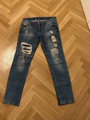 Blå slitna jeans med hål och patchar - Jeansen har en smal passform och detaljer som färgstänk och dekorativa sömmar. Perfekta för en edgy streetstyle-look.