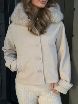 Vårjacka Gina Tricot - Stilren beige kappa med stor fluffig fuskpäls-krage och knäppning framtill. Jackan har raka ärmar, fickor på sidorna och är tillverkad i ett mjukt, ull-liknande material. Perfekt för dig som vill ha en trendig och mysig look. Passar bra nu till våren.