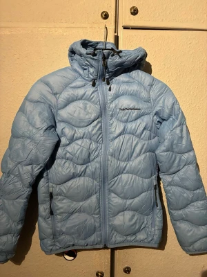 Ljusblå dunjacka från Peak Performance - Snygg ljusblå dunjacka från Peak Performance med huva och dragkedja framtill. Jackan har två sidofickor med dragkedja och quiltat mönster. Perfekt nu för våren och riktigt skön att bära. Klassisk logga på bröstet. Se sista bilden så finns de en liten fläck men den syns knappt, därför sänkte jag priset lite.
