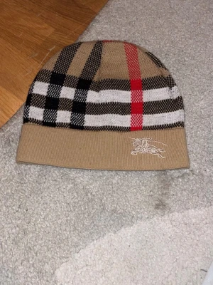 Beige Burberry-mössa med rutmönster - Säljer en beige mössa från Burberry med det klassiska rutmönstret i svart, vitt och rött. Mössan har en vikt kant och en broderad logga på sidan. Perfekt accessoar för dig som vill ha en stilren och ikonisk look.