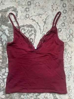 Vinröd linnetopp med spetskant - Snygg vinröd linnetopp med tunna axelband och v-ringning dekorerad med spets. Toppen har en croppad passform och är tillverkad i mjukt material som känns skönt mot huden. Perfekt att styla med jeans eller kjol. Passar S/M