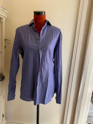 Blå randig skjorta med knappar - Klassisk blå skjorta med tunna vita ränder och knäppning framtill. Skjortan har lång ärm och en avslappnad passform, perfekt för en stilren look. Tillverkad i ett lätt och bekvämt bomullsmaterial.