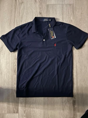 Marinblå piké från Polo Ralph Lauren - Klassisk marinblå pikétröja från Polo Ralph Lauren med korta ärmar och röd broderad logga på bröstet. Tröjan har krage och knappar framtill, tillverkad i mjuk bomull som känns skön mot huden. Perfekt för en stilren och avslappnad look.