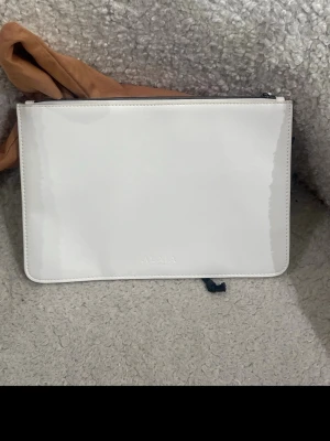 Vit kuvertväska i lack från Alaïa - Snygg vit kuvertväska från Alaïa i glansigt lackmaterial. Väskan har en stilren, rektangulär form och dragkedja upptill. Insidan är ljusbeige och slät, perfekt för att bära det viktigaste. Diskret Alaïa-logga framtill.