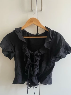 Svart croppad blus med snörning - Trendig svart croppad blus med volangkant och snörning framtill. Korta ärmar och v-ringad design ger en snygg och ungdomlig look. Perfekt att styla med jeans eller kjol för en cool outfit.