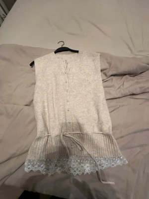 Beige stickad väst med spetskant - Snygg beige stickad väst med knappar framtill och dekorativ snörning i midjan. Nederkanten har en fin spetsdetalj som ger en romantisk touch. Perfekt att styla över en skjorta eller t-shirt för en trendig lager-på-lager-look.