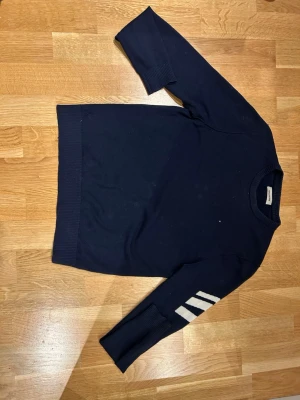 Mörkblå stickad tröja Zadig & Voltaire - Mörkblå stickad tröja från Zadig & Voltaire med rund halsringning och ribbade muddar. Ena ärmen har tre vita ränder som detalj. Perfekt för dig som gillar stilrena och sportiga plagg.