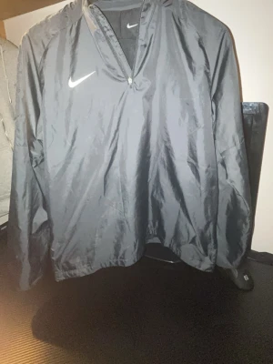 Svart Nike vindjacka med dragkedja - Svart vindjacka från Nike med halv dragkedja framtill och klassisk vit swoosh-logga på bröstet. Jackan har en lös passform och är tillverkad i lätt, glansigt material som skyddar mot vind. Perfekt för sport eller chill dagar.
