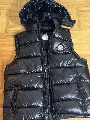 Svart dunväst från Moncler med huva - Snygg svart dunväst från Moncler med glansig finish och avtagbar huva. Västen har två dragkedjefickor framtill och klassisk Moncler-logga på bröstet. Fylld med vitt gåsdun för extra värme. Perfekt för lager-på-lager-stil.