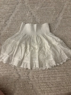 Vit volangkjol med spetsdetaljer  - Supersöt vit kortkjol med bred smock i midjan som kan vikas ner och flera lager volanger. Kjolen har fina spetsdetaljer och broderade mönster längs kanten. kjolen har även en innerkjol vilket gör att den inte blir så genomskinlig. Aldrig använd endast testad men lappen har åkt av. Den finns inte längre att köpa. Från Neo Noir 