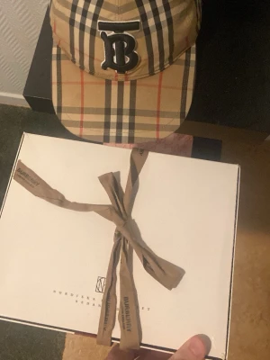 Burberry beige rutmönstrad keps - Säljer en klassisk Burberry keps i beige med det ikoniska rutmönstret och en stor svart-vit 'B' broderad framtill. Kepsen har böjd skärm och justerbar rem bak. Tillverkad i bomull och har svart insida. Perfekt för dig som vill ha en stilren och trendig accessoar.