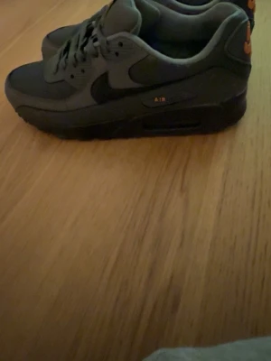 Nike Air Max 90 mörkgrå sneakers - Snygga Nike Air Max 90 sneakers i mörkgrått med svarta detaljer och orange logga på plösen och hälen. Klassisk chunky sula med synlig Air-enhet och robust design. Perfekta för dig som gillar streetwear och sportig stil.
