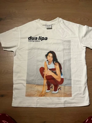 Vit Dua Lipa t-shirt från Bershka - Cool vit t-shirt från Bershka med stort tryck av Dua Lipa och texten 'training season' på bröstet. Klassisk rund hals och korta ärmar, perfekt för dig som gillar musik och streetstyle. Materialet är mjuk bomull och trycket är färgstarkt och tydligt.