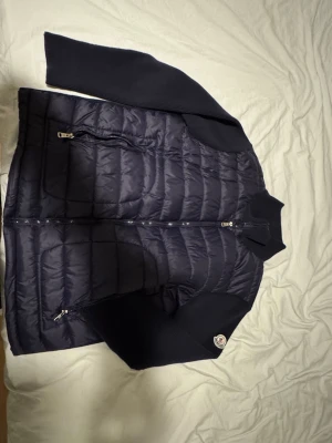 Mörkblå dunjacka från Moncler - Snygg mörkblå dunjacka från Moncler med quiltad front och stickade ärmar. Jackan har två dragkedjeförsedda fickor och en hög krage. Klassisk Moncler-logga på ena ärmen. Materialmix med dunfylld kropp och ribbstickade detaljer ger en modern look. As bra fynd nu inför säsongen för dig som vill klä sig stilfullt. Storlek M men passar perfekt på L också pris kan prutas vid snabba köp, inga orimliga prisförslag!