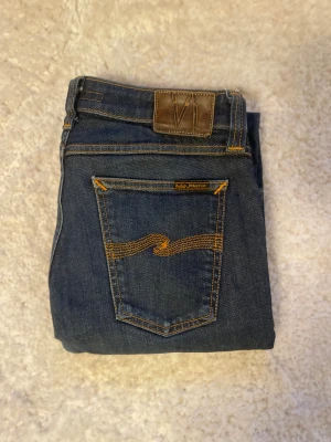 Mörkblå jeans från Nudie Jeans - Snygga mörkblå jeans från Nudie Jeans med klassisk femficksdesign och kontrastsömmar i orange. Modellen är slim och är tillverkad i slitstarkt denim med Nudie-logga på bakfickan och läderpatch i midjan. Perfekt för dig som gillar stilrena och tidlösa jeans. Är egentligen w30 men är som en w28