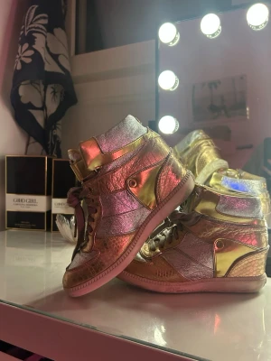 Kilklackar från Michael Kors - Säljer ett par riktigt snygga high top sneakers från Michael Kors i metallic guld och rosa. Skorna har coola detaljer, snörning och MK-logga framtill. Materialet är glansigt och skimrande, vilket gör dem till ett statement-piece för dig som vill sticka ut. 