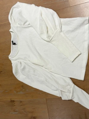 Vit långärmad topp från Vero Moda - Trendig vit långärmad topp från Vero Moda med puffiga ärmar och ribbad struktur. Toppen har rund halsringning och är tillverkad i ett mjukt material som känns skönt mot huden. Perfekt för dig som gillar stilrena och moderna plagg.