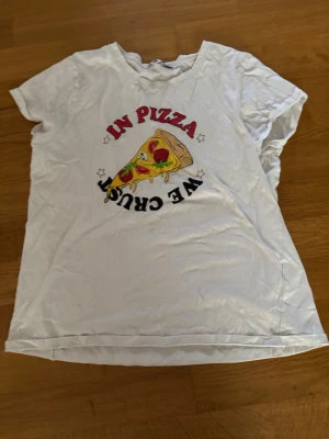 Vit pizza-tshirt från Sister - Cool vit t-shirt från Sister med ett färgglatt pizzatryck och texten 'In Pizza We Crust' på bröstet. T-shirten har korta ärmar och är gjord i mjuk bomull, perfekt för dig som älskar pizza och vill ha en chill look.
