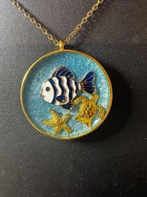 Halsband med fisk och havsmotiv - Unikt halsband med runt hänge i blått och guld. Hänget har ett detaljerat motiv med en blåvit fisk, en guldfärgad sjöstjärna och en guldfärgad sköldpadda. Kedjan är tunn och guldfärgad. Perfekt för dig som gillar havsinspirerade accessoarer.