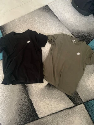 Nike T-shirts svart & olivgrön XL - Två stilrena Nike T-shirts i svart och olivgrönt med klassisk logga på bröstet. Båda är kortärmade och tillverkade i mjuk bomull, perfekta för en avslappnad och sportig look. Säljes tillsammans, passar dig som gillar enkel och snygg streetwear.