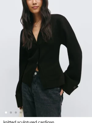 Svart knappad kofta med vida ärmar - Stilren svart kofta med stora knappar framtill och vida ärmar. Koftan har en djup v-ringning och är tillverkad i ett mjukt tyg som ger en avslappnad look. Perfekt att matcha med jeans eller kjol för en trendig outfit.