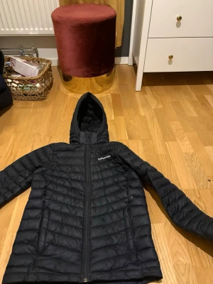 Svart dunjacka med huva från Peak Performance - Snygg svart dunjacka från Peak Performance med huva och dragkedja framtill. Jackan har två sidofickor med dragkedja och diskret logga på bröstet. Perfekt för kalla vinterdagar och har en stilren, sportig look.                                                          Ordinarie pris: ca 1500