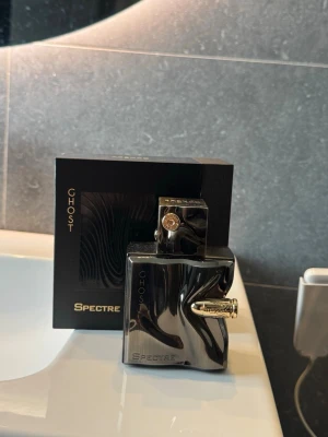 Ghost Spectre parfym svart flaska - Ghost Spectre parfym i en unik, svart och metallisk flaska med vågig design och gulddetaljer. Flaskan har en modern, kantig form och kommer i en matchande svart förpackning med guldtext. Perfekt för dig som gillar exklusiva och stilrena dofter.