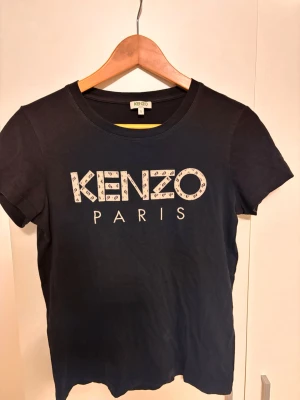 Svart Kenzo Paris t-shirt - Snygg svart t-shirt från Kenzo Paris med stort logotryck framtill. Klassisk rund halsringning och korta ärmar. Tillverkad i mjuk bomull som känns skön mot huden. Perfekt för dig som gillar stilrena och trendiga plagg med designer-touch.