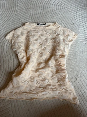 Beige topp med 3D-mönster från Bik Bok - Säljer en beige topp från Gina tricot med coolt 3D-mönster över hela tyget. Toppen är kortärmad och har en något croppad passform som är perfekt till höga jeans eller kjol. Materialet känns lätt och luftigt, perfekt för varma dagar.