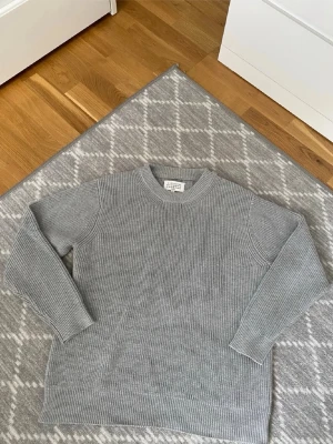 Grå stickad tröja Maison Margiela - Säljer en stilren grå stickad tröja från Maison Margiela. Tröjan har rund halsringning, ribbstickade muddar och är tillverkad i mjukt ullmaterial. Klassisk rak passform och diskreta detaljer i nacken. Perfekt för lager på lager.m/L