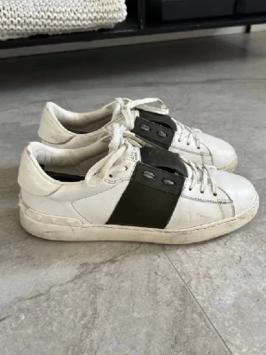 Valentino skor  - Snygga vita sneakers från valentino med en bred svart läderdetalj på sidan och vita skosnören. Klassisk låg modell med rund tå och diskret branding på hälen. Perfekta för en stilren och trendig look. Hör av dig vid minsta intresse!⭐️✨