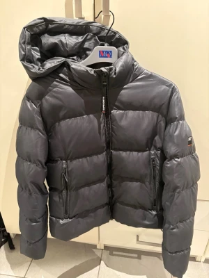Mörkgrå pufferjacka från Superdry - Super snygg mörkgrå ( lite glansigare) pufferjacka från Superdry med huva och två stora dragkedjeförsedda fickor framtill. Jackan har en hög krage med en bekväm luva. Perfekt för kalla vinterdagar och riktigt trendig stil. Jackan är i nyskick, näst intill oanvänd😍🥰❤️