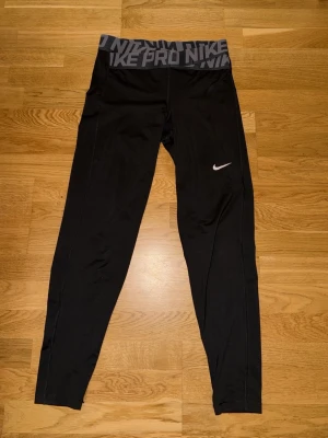 Svarta Nike Pro Dri-FIT leggings - Säljer ett par svarta Nike Pro Dri-FIT leggings med bred midjeresår där det står 'NIKE PRO' i grått. De har en vit Nike-logga på benet och är tillverkade i ett stretchigt funktionsmaterial som andas. Perfekta för träning eller chill.