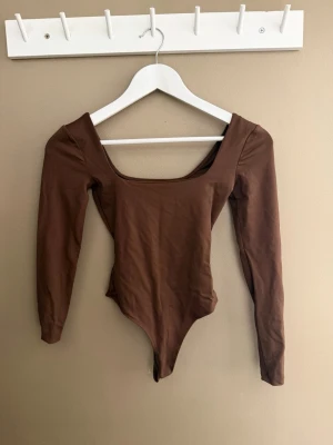 Brun långärmad body från Zara - Snygg brun body från Zara med långa ärmar och bred, fyrkantig urringning. Tillverkad i mjukt stretchigt material som sitter tight och bekvämt. Perfekt att styla med jeans eller kjol för en trendig look.