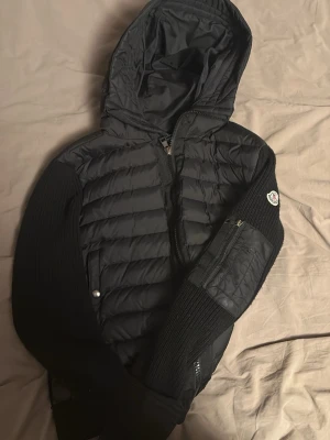 Svart pufferjacka från Moncler - Snygg svart pufferjacka från Moncler med stickade ärmar och detaljer. Jackan har quiltad front och huva, dragkedja framtill och en ficka med dragkedja på ärmen. Moncler-logga på vänster ärm. Perfekt för kyliga dagar.